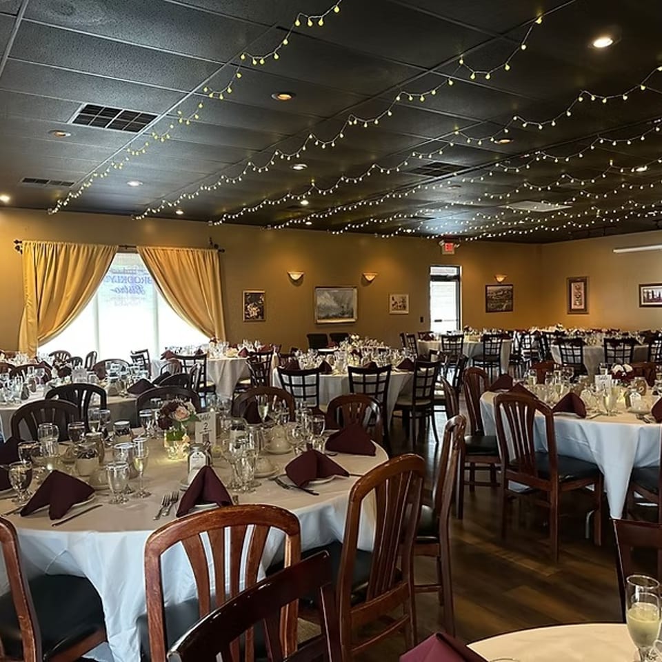 A restaurant dining room with round tables set with white tablecloths, maroon napkins, and string lights.