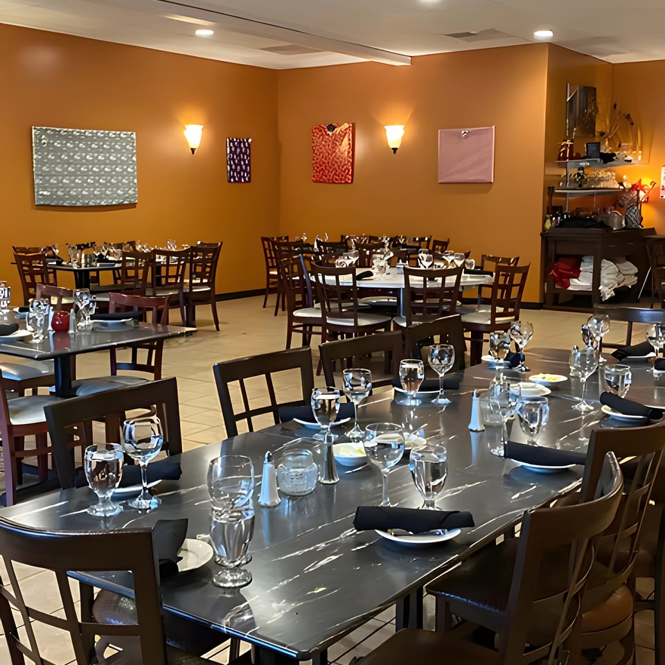 Restaurant dining room with multiple tables set with glasses and black napkins, orange walls, and modern art.